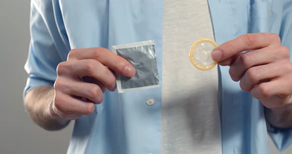 Close Up of Man Holding Unwrapped Condom in Hand Standing Isolated on Grey Background