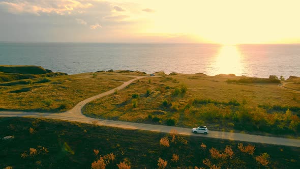 Above View of White Car Riding Country Road Turning to Sea Coast at Warm Sunset