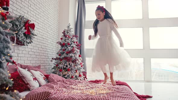 Cute Girl in White Dress Jumping on Bed with Pillow Christmas Morning Time Kids Party