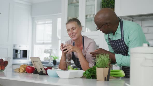 Smiling senior diverse couple wearing blue aprons and drinking wine in kitchen