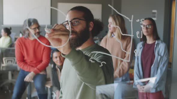 Young Man Drawing on Glass Board and Talking to Business Team