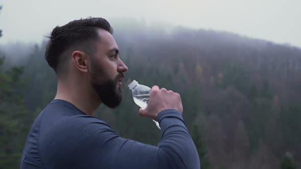 Side View of Strong Man with Big Cool Beard Drinking Water Outside Against Foggy Forest