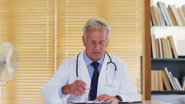 Portrait of male caucasian doctor of talking and waving looking at camera while sitting in hospital