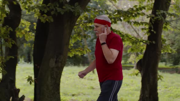 Male Senior Person Running Along the Road in Park. Mature Runner Man Training, Listening Music