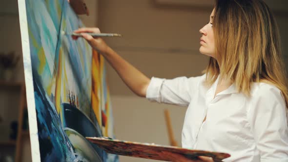 Attractive Young Woman in White Shirt Is Painting Seascape in Oil on Canvas Holding Brush and Color