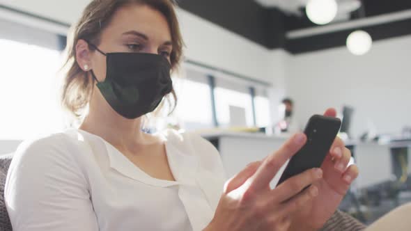 Caucasian businesswoman wearing face mask and using smartphone in office