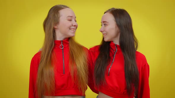 Cheerful Young Female Twins Looking at Each Other Turning to Camera Smiling Gesturing Forefinger Up