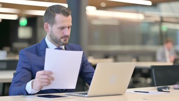 Middle Aged Businessman Reading Documents While Using Laptop