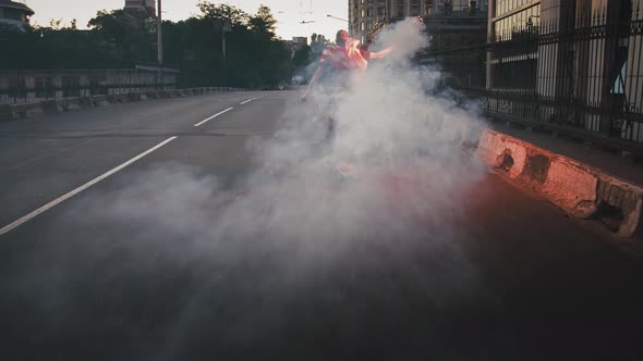 Young Guy in Flag of USA Tied on His Neck is Skateboarding Along Street with Burning Red Signal