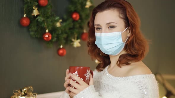Sad Young Woman in Medical Mask Holds Red Christmas Cup of Tea