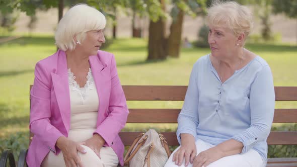 Nice Mature Caucasian Women Sitting at the Bench in the Summer Park. Senior Woman with Blond Hair