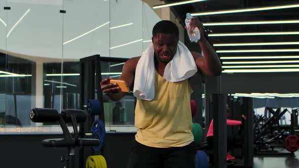 Black Man Looks at Phone in Gym While Drinks Water While Relaxing