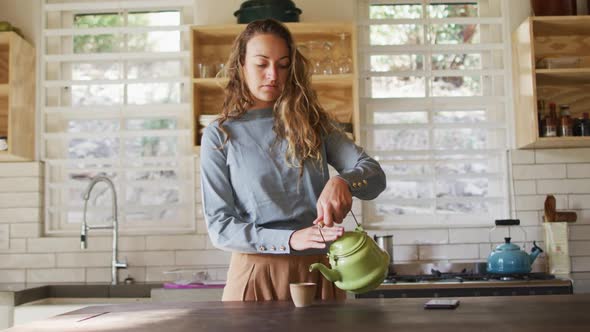 Happy caucasian woman standing at counter in cottage kitchen pouring tea from teapot and smiling