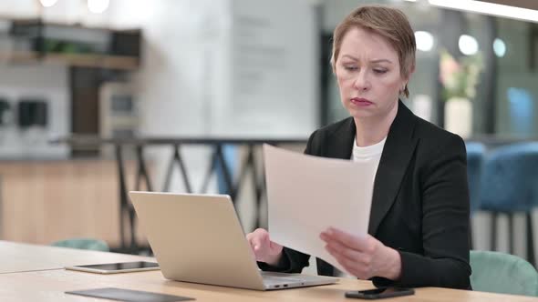 Old Businesswoman Working on Laptop with Documents in Office