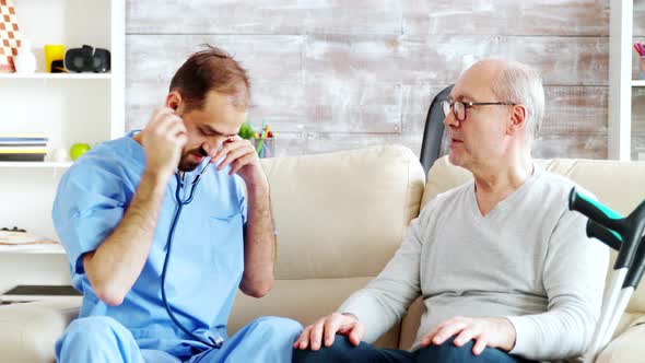 Revealing Shot of Young Male Nurse Listening To Old Retired Man Heartbeat