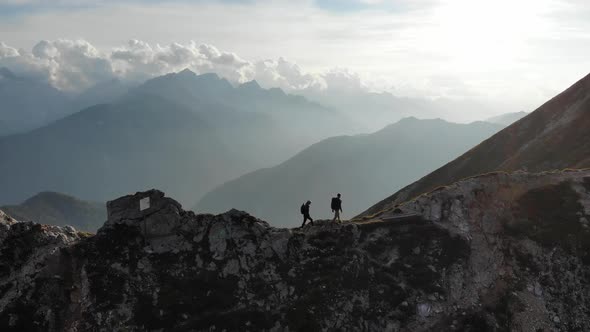 Drone Flight Over Two People Hiking On Mountain Top In Front Of Silhouette Of The Alps