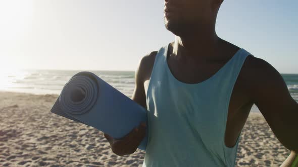 Smiling african american man holding yoga mat, standing on beach by the sea