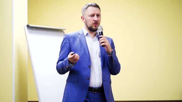 Bearded businessman in suit with microphone gives the lecture indoors.