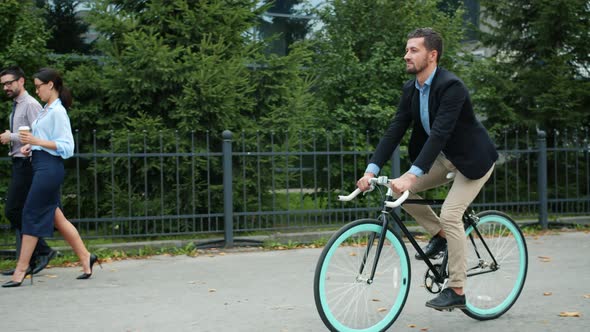 Good-looking Man Riding Bike Wearing Formalwear While Businesspeople Walking By