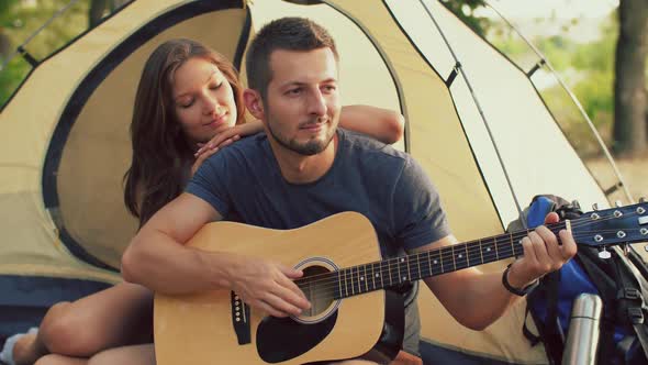 Man Playing Guitar for His Girlfriend Sitting at the Camping Tent