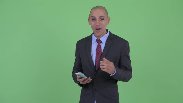 Happy Bald Multi Ethnic Businessman Using Phone and Looking Surprised