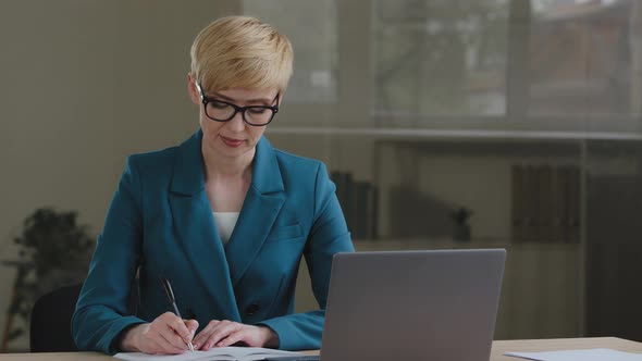 Middle Aged Female Entrepreneur Wears Glasses Using Laptop Writing Notes Sitting at Office Table