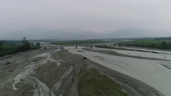 Aerial view of bridge over riverbed swollen by rushing water of rainy season, misty horizon of mount