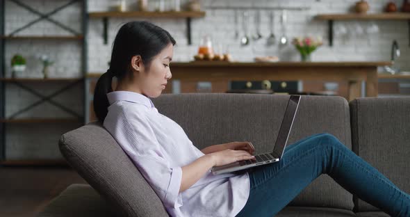 Asian Woman Resting on Couch Using Laptop Notebook Looking at Screen Typing Message at Living Room