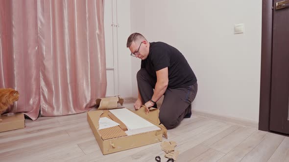 a Young Man with Glasses Opens a Large Box on the Floor Tearing Open Cardboard