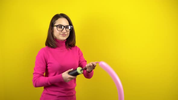 A Young Woman Inflates a Long Pink Balloon with a Pump on a Yellow Background. Girl in a Pink