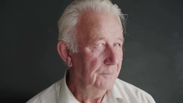 Portrait of Senior Man Turns and Looks with Light Smile at Camera on Background