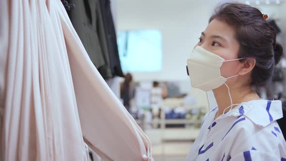 asian female shopper wearing facial virus protective mask choosing clothes at the store