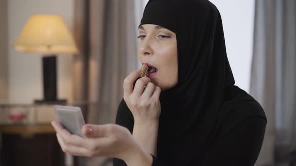 Portrait of Young Beautiful Muslim Lady Looking at Mirror and Applying Lipstick. Modern Woman in