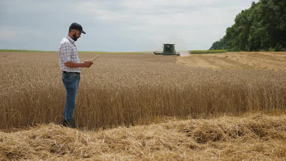 A Farmer Uses a Digital Tablet, in the Background Is a Harvester