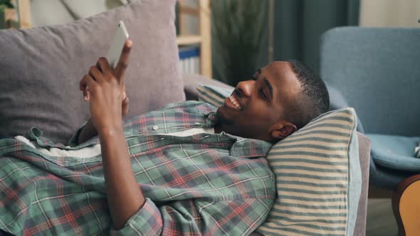 Joyful African American Man Is Holding Smartphone, Touching Screen and Smiling Lying on Couch at