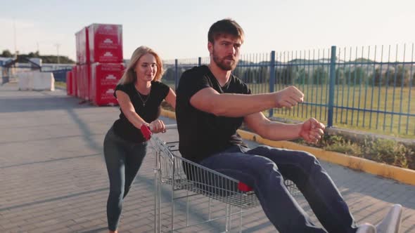 Young Woman Rolls Attractive Guy in the Trolley on Parking Lot of the Supermarket at Sunset