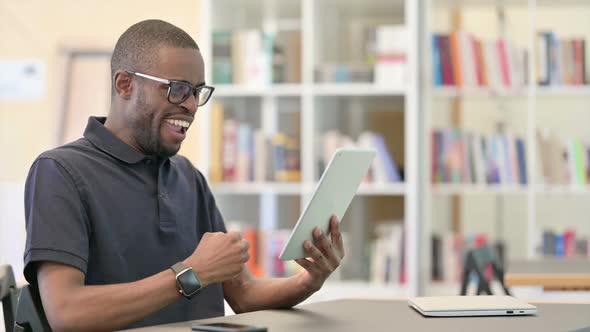 Success Young African Man Celebrating on Tablet in Library