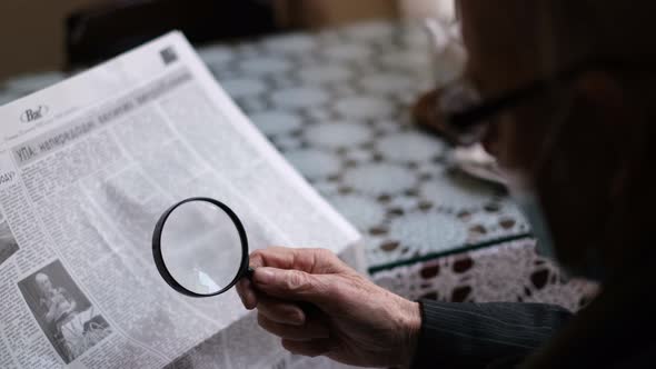 An Old Pensioner with Glasses is Sitting in a Cafe and Reading a Newspaper