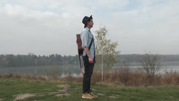 Hippie Man Playing an Acoustic Guitar in the Autumn Forest and Lake