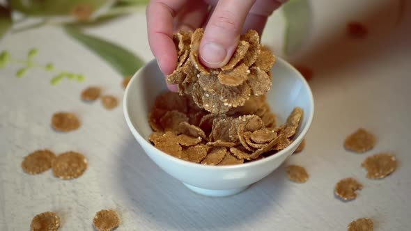 Female Hand Scatters Pours Out Throws Corn Flakes Into a White Bowl