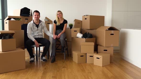 A Happy Moving Couple Sits on Chairs in an Empty Apartment and Smiles at Camera in Empty Apartment