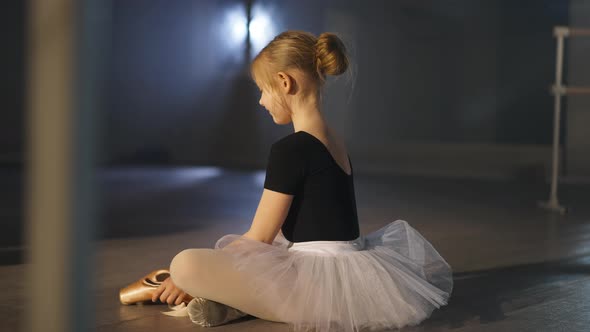 Side View Wide Shot of Happy Caucasian Girl in Tutu Smiling Admiring Golden Pointes Sitting on Floor