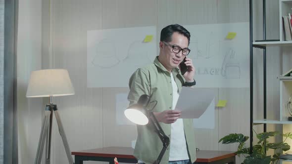 Asian Man Designer Talking On Smartphone While Looking At The Layout Bond In Hand And Walking