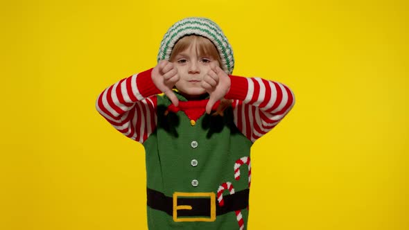 Displesed Kid Girl in Christmas Elf Santa Helper Costume Keeps Thumb Down and Shows Dislike Gesture