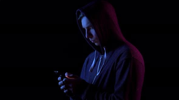 A Young Man in a Hood with a Phone in His Hands. Hacker Makes a Hack Through the Phone. Blue and Red