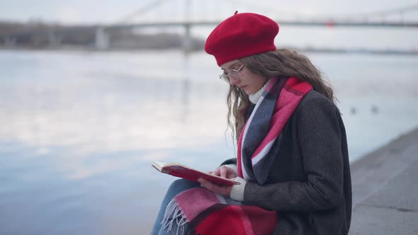 Side View of Intelligent Confident Beautiful Young Woman Reading Book Sitting on Urban Embankment on