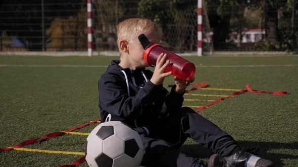 Little Tired Boy Sitting on the Football Field and Drinking Water From a Bottle