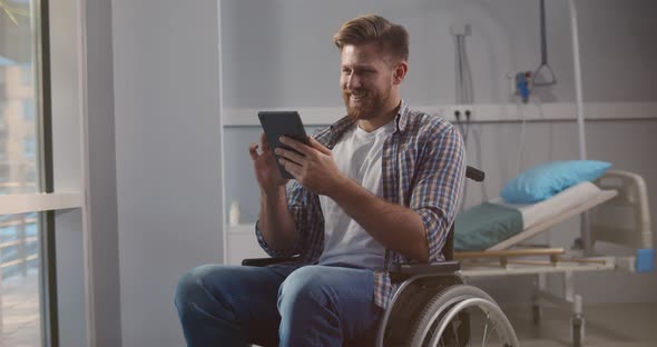 Disabled Young Caucasian Man Sitting in Wheelchair and Using Digital Tablet