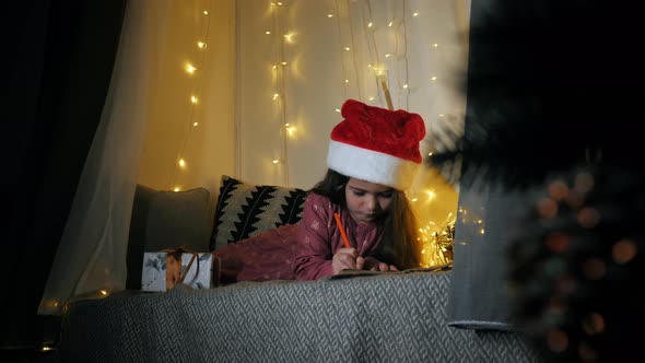 Little Girl in Santa Hat Is Writing a Letter To Santa Claus on Windowsill Decorated for Christmas