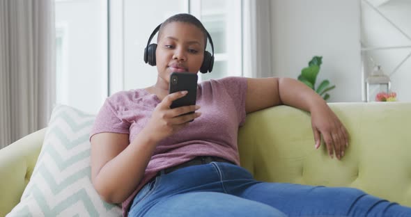 Happy african american plus size woman sitting on sofa, wearing headphones and using smartphone